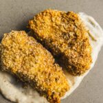 Crispy breaded chicken breasts with creamy herb sauce on neutral background.