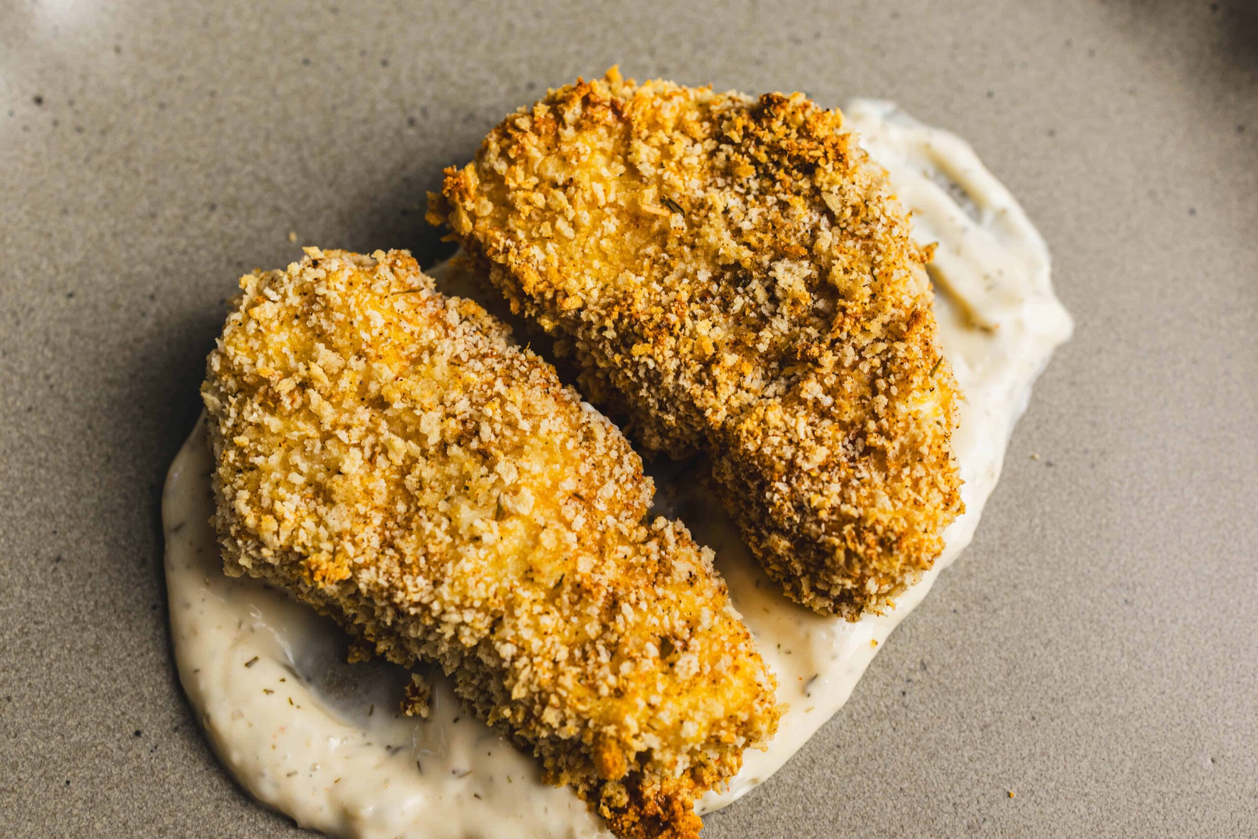 Crispy breaded chicken breasts with creamy herb sauce on neutral background.
