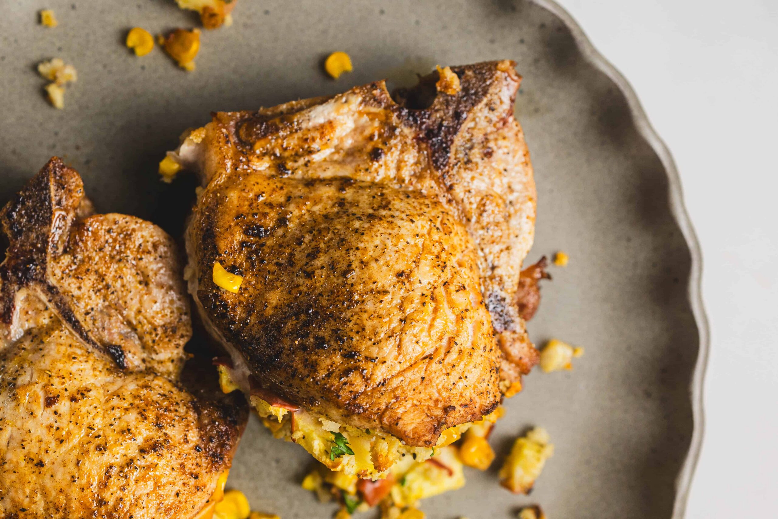 Roasted chicken thighs with crispy skin served on a rustic grey plate, paired with colorful corn and vegetable stuffing, perfect for a delicious homemade meal.