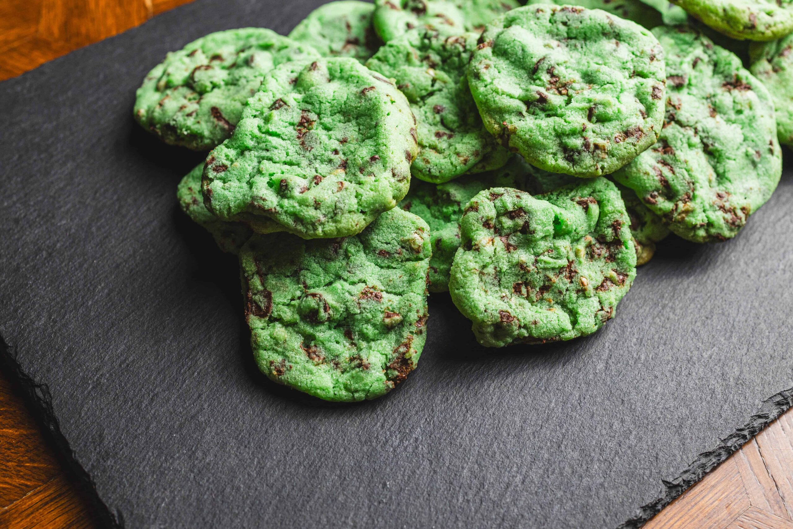 Vibrant green mint chocolate chip cookies on a dark slate serving platter, showcasing homemade baked treats with colorful chocolate chunks, perfect for dessert or snack.