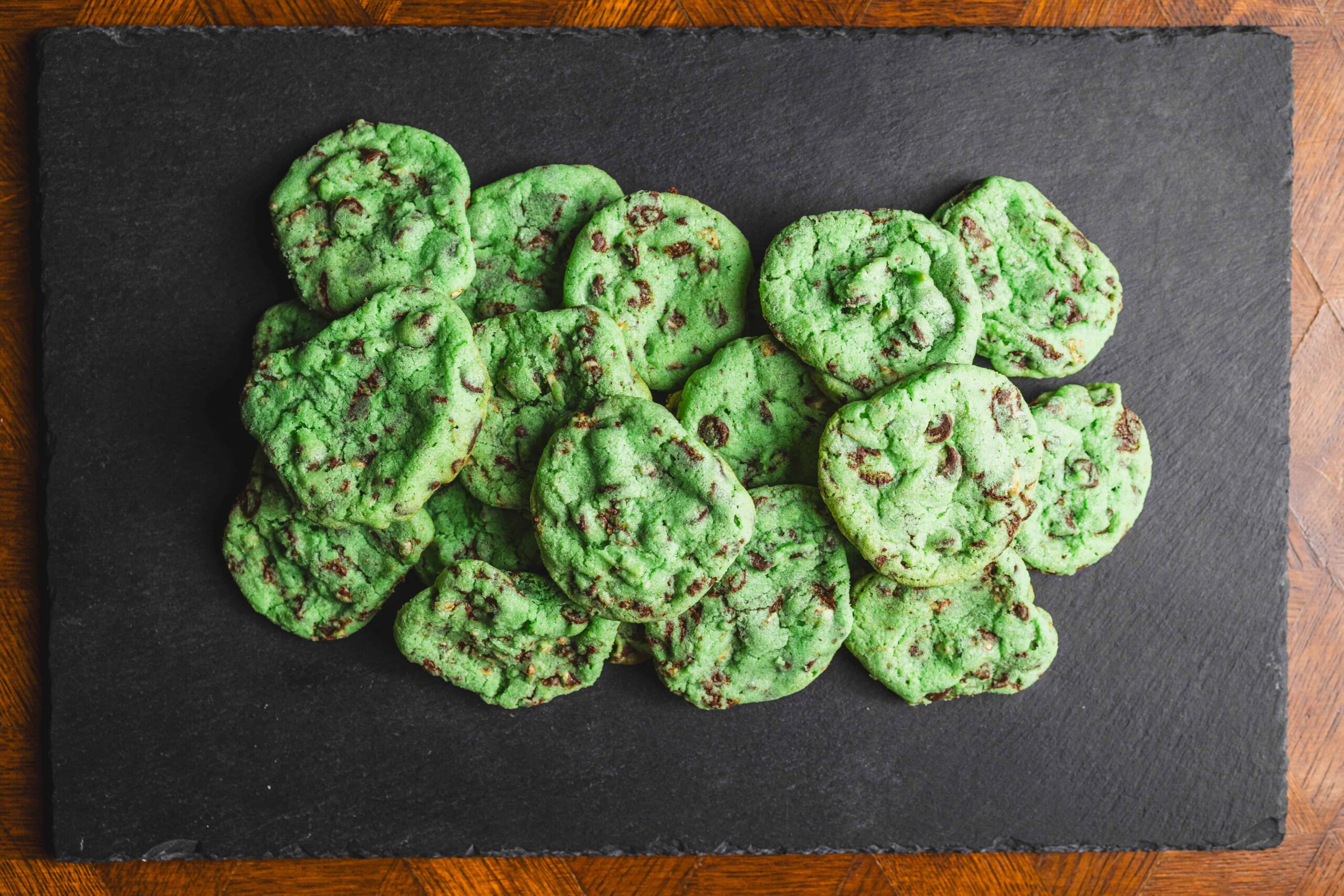 Fresh green mint chocolate chip cookies arranged on a black slate serving board, showcasing vibrant color and chocolate chunks, perfect for mint chocolate lovers and cookie recipes.