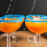 Bright orange cocktails in classic coupe glasses with blue sugar rim decorations and a citrus garnish, served on a wooden tray, perfect for festive celebrations or summer parties.