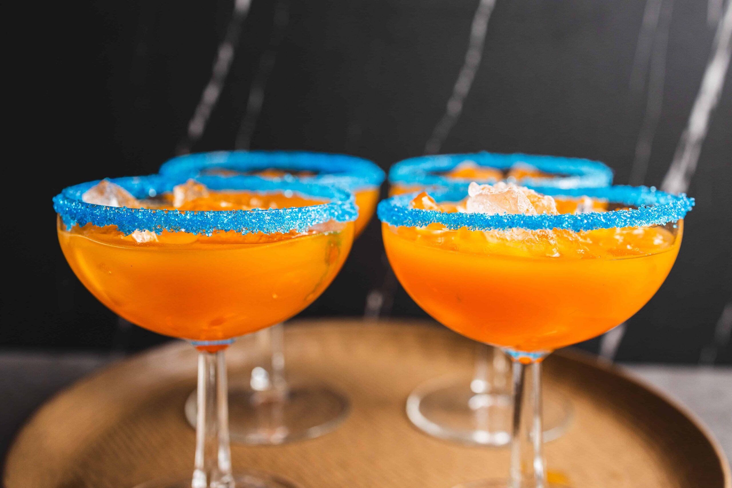 Bright orange cocktails in classic coupe glasses with blue sugar rim decorations and a citrus garnish, served on a wooden tray, perfect for festive celebrations or summer parties.