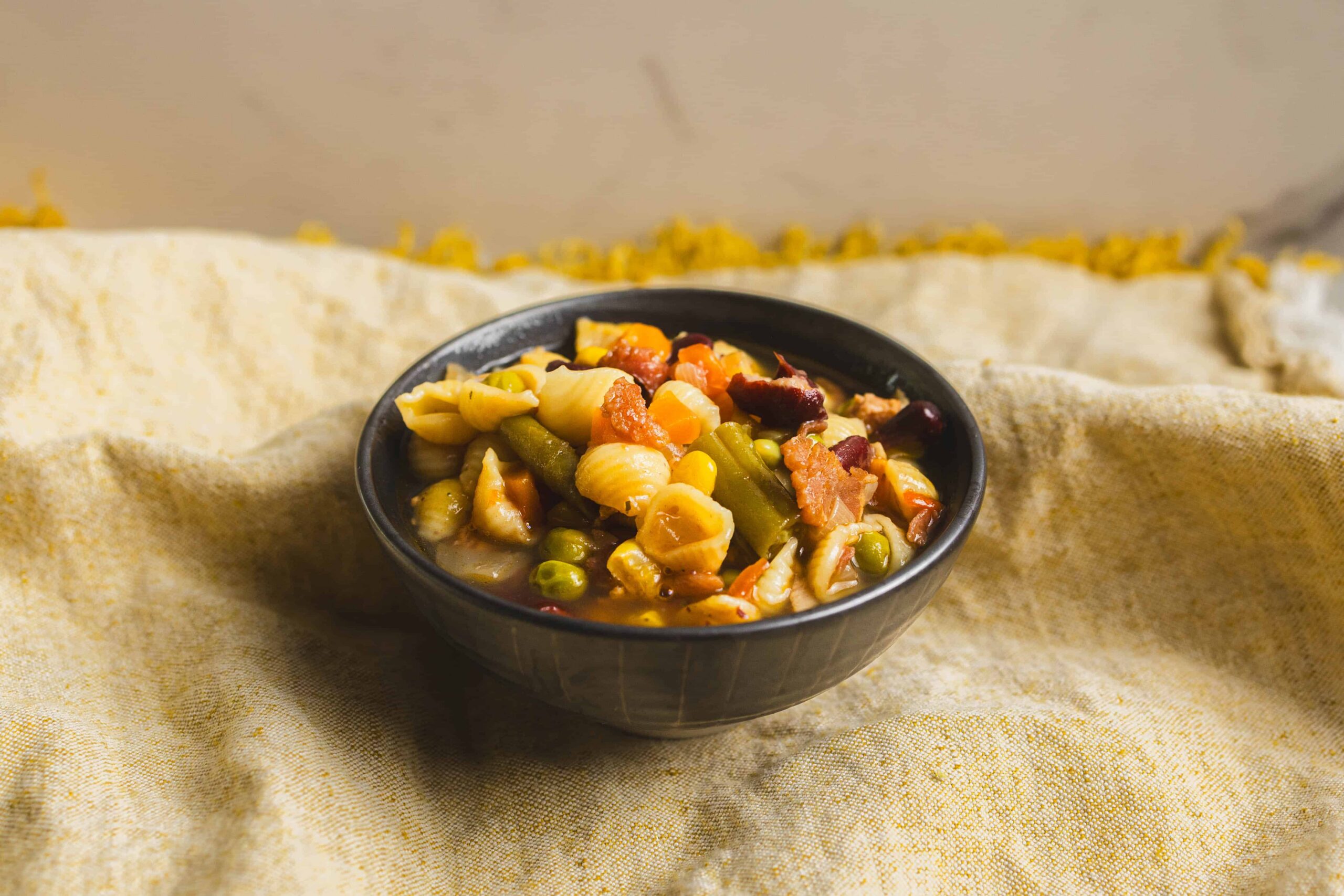 Assorted vegetable pasta salad in a black bowl on a textured beige cloth, featuring shell pasta, green beans, peas, corn, and diced vegetables. Perfect for healthy, easy-to-make vegetarian meals and side dishes.