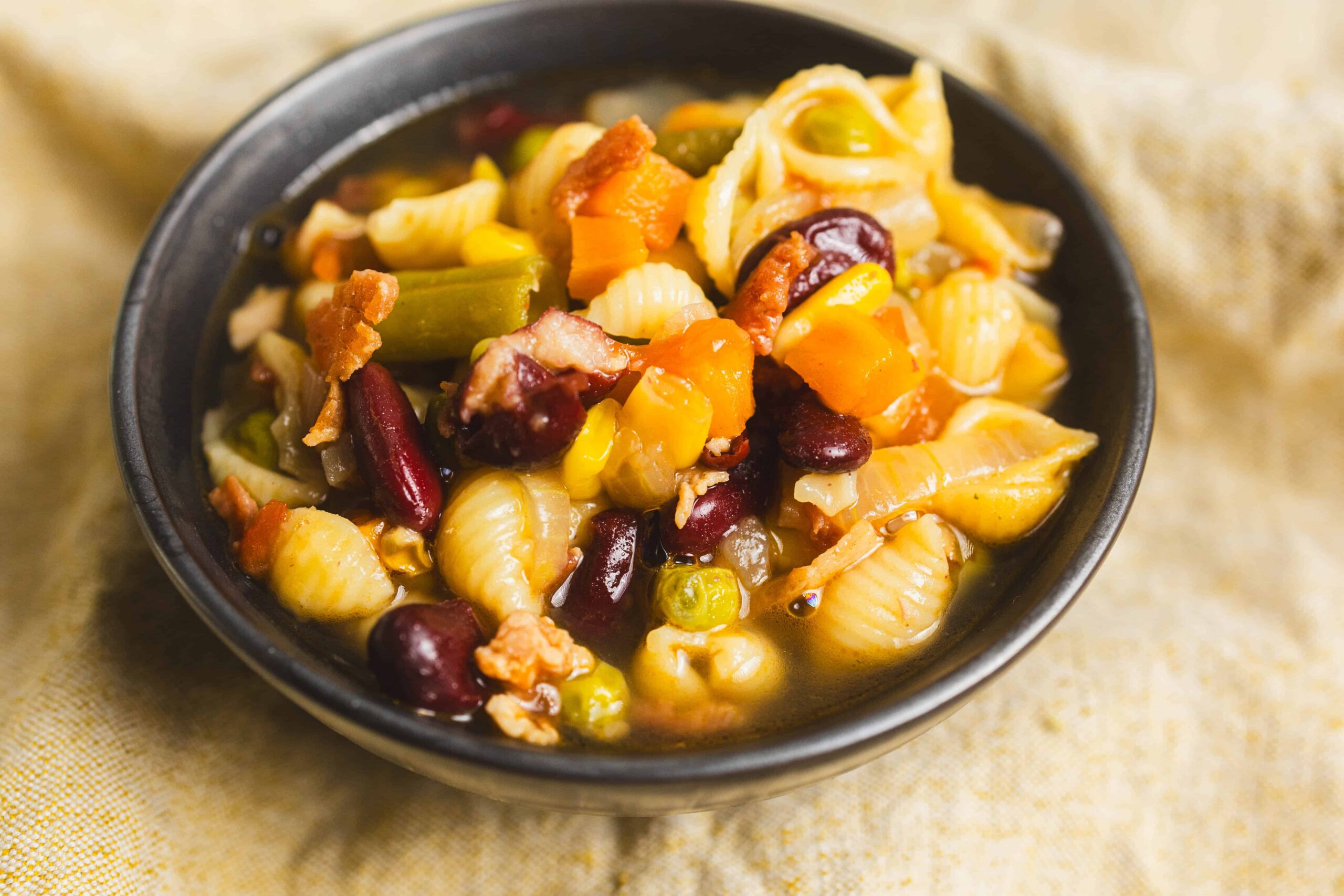 Juicy mixed vegetable pasta salad with beans, corn, and diced vegetables in a black bowl on beige fabric. A flavorful, colorful vegetarian dish perfect for lunch or dinner.