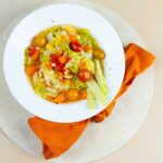 Fresh vegetable soup with cherry tomatoes, carrots, celery, and lettuce served in a white bowl on a beige table with an orange napkin.