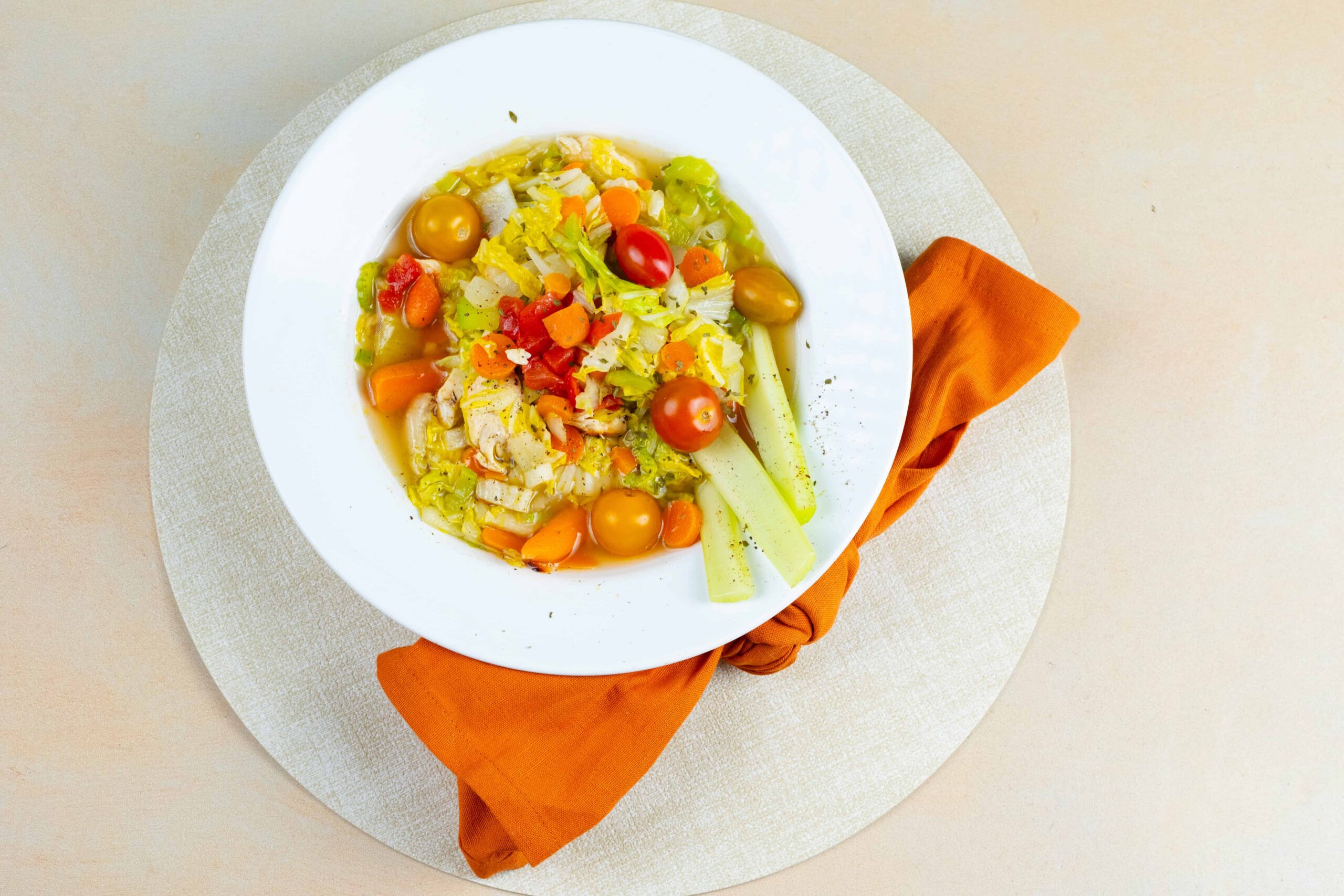 Fresh vegetable soup with cherry tomatoes, carrots, celery, and lettuce served in a white bowl on a beige table with an orange napkin.