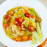 Vibrant vegetable chicken soup with cherry tomatoes, carrots, lettuce, and celery in a clear broth, served on a white plate with a burnt orange napkin for a healthy, colorful meal.