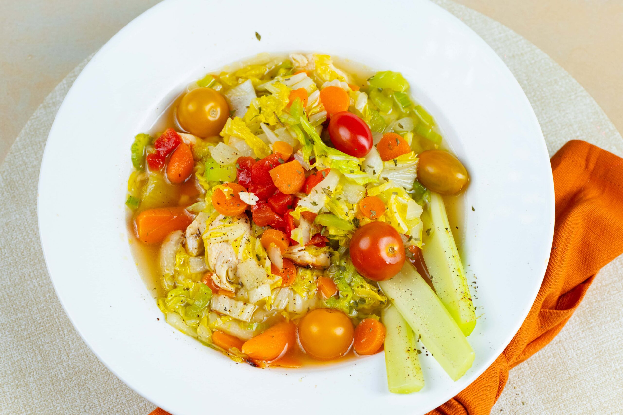 Vibrant vegetable chicken soup with cherry tomatoes, carrots, lettuce, and celery in a clear broth, served on a white plate with a burnt orange napkin for a healthy, colorful meal.