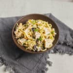 Creamy chicken and mushroom pasta in a brown bowl, garnished with herbs, served on a rustic gray cloth. Perfect for easy dinner recipes and comfort food lovers.