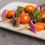 Skewers with cherry tomatoes, red onion, beef, and green herbs on a white plate. Fresh, colorful, and delicious-looking grilled meat skewers perfect for summer recipes and outdoor barbecues.