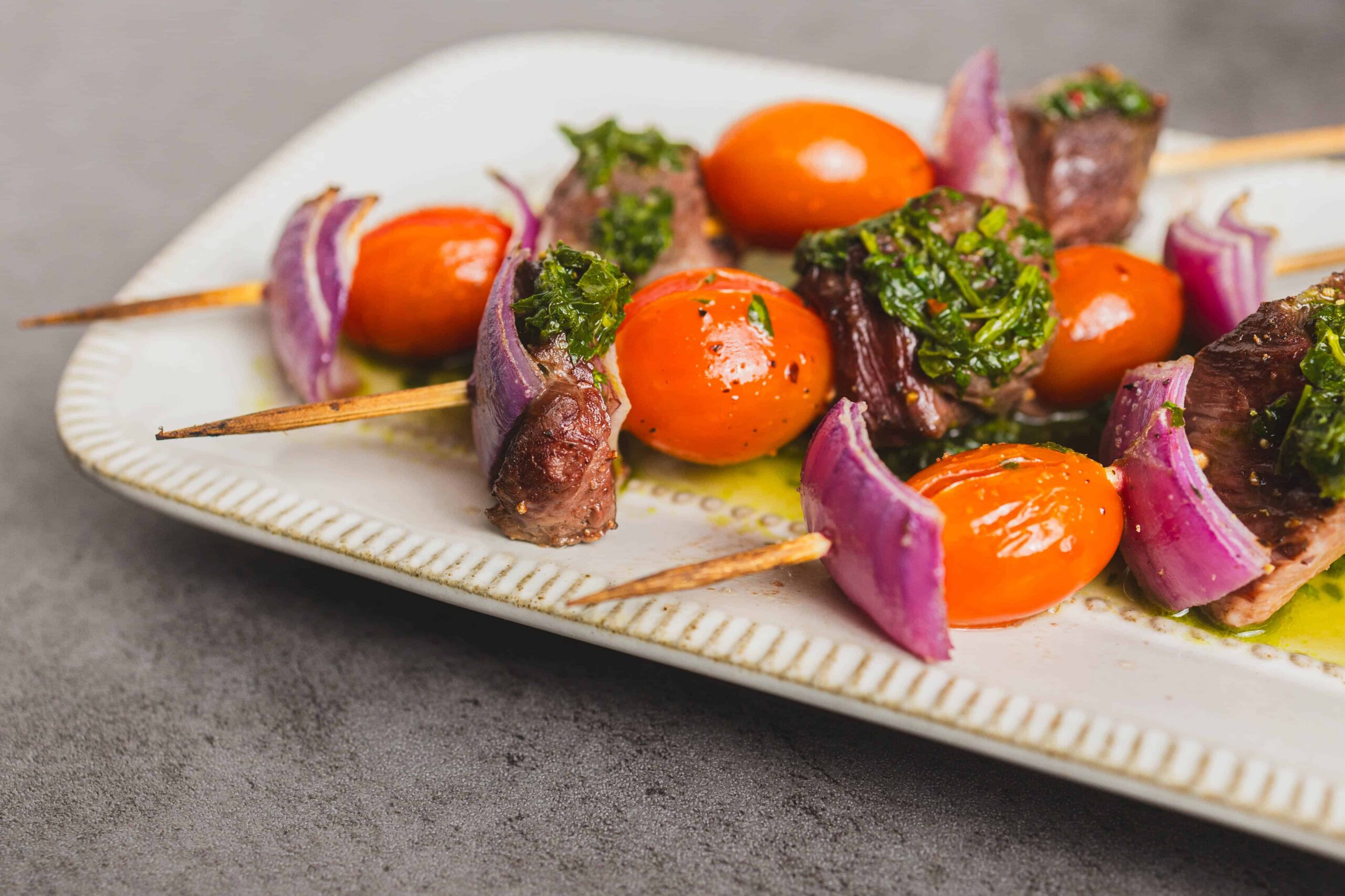 Skewers with cherry tomatoes, red onion, beef, and green herbs on a white plate. Fresh, colorful, and delicious-looking grilled meat skewers perfect for summer recipes and outdoor barbecues.