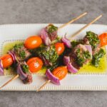 Grilled beef skewers with cherry tomatoes, red onion, and herb marinade served on a white plate, garnished with green sauce, perfect for summer barbecue recipes.