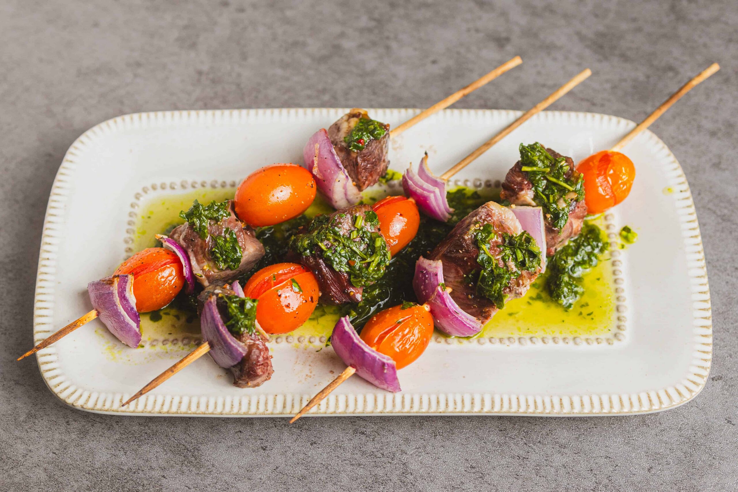 Grilled beef skewers with cherry tomatoes, red onion, and herb marinade served on a white plate, garnished with green sauce, perfect for summer barbecue recipes.