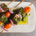 Skewers of grilled beef, cherry tomatoes, red onions, and green herbs served on a white plate with herb marinade and oil drizzle, perfect for tasty barbecue recipes.