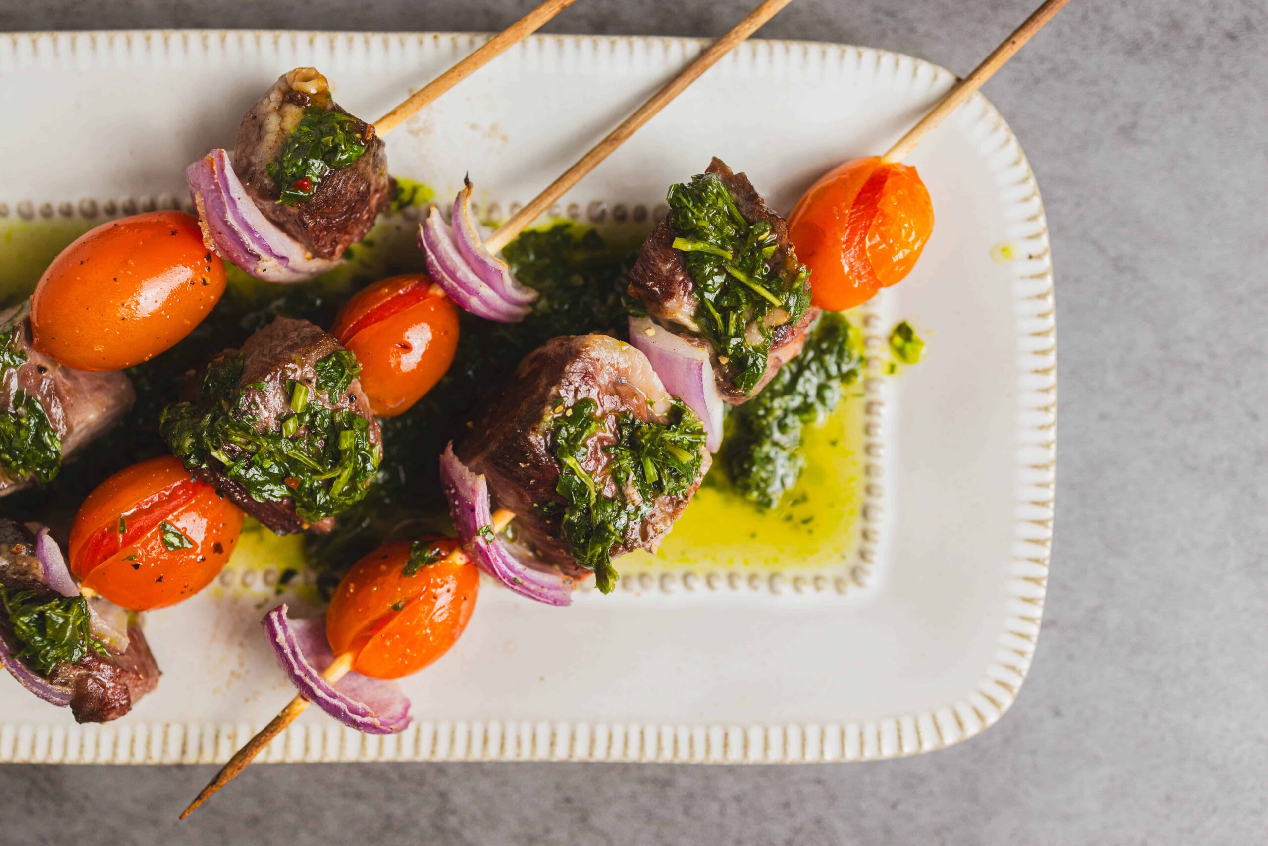 Skewers of grilled beef, cherry tomatoes, red onions, and green herbs served on a white plate with herb marinade and oil drizzle, perfect for tasty barbecue recipes.