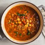 Hearty bowl of homemade vegetable and ground meat chili soup in a rustic enamel pot, garnished with diced corn and fresh herbs, served on a light wooden surface with a cozy woven towel.
