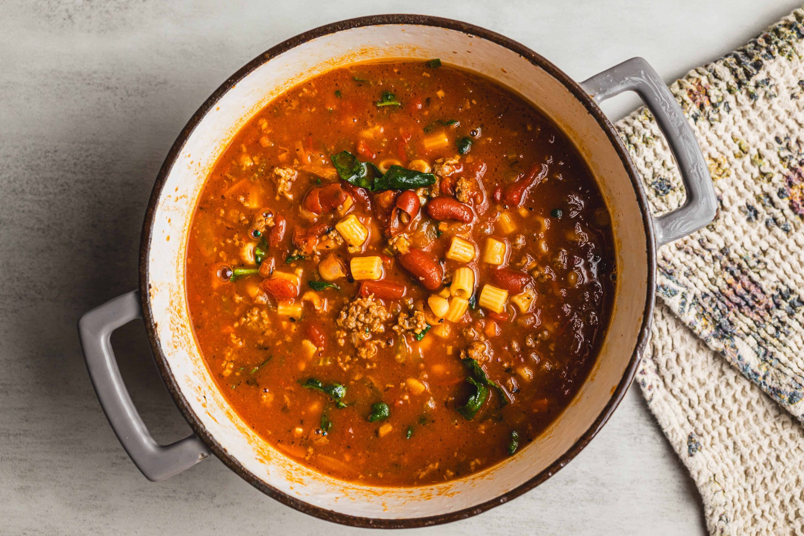 Hearty bowl of homemade vegetable and ground meat chili soup in a rustic enamel pot, garnished with diced corn and fresh herbs, served on a light wooden surface with a cozy woven towel.