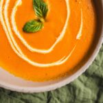 Creamy pumpkin soup garnished with basil leaves, served in a white bowl on a green textured cloth. Perfect for fall, vegan, and hearty comfort food.