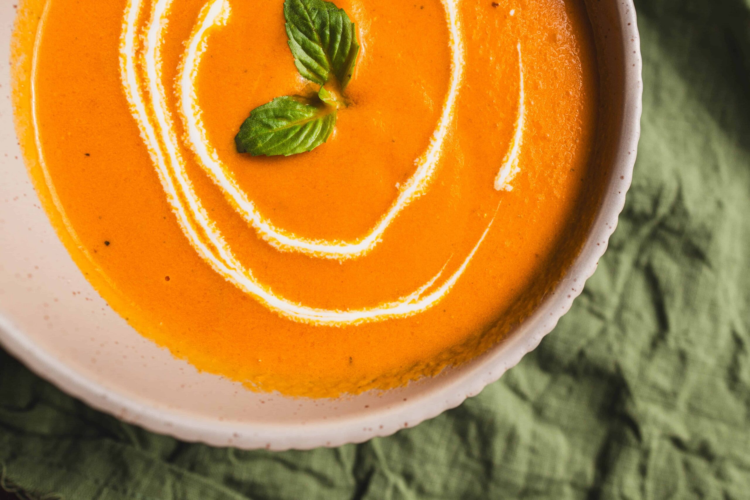 Creamy pumpkin soup garnished with basil leaves, served in a white bowl on a green textured cloth. Perfect for fall, vegan, and hearty comfort food.