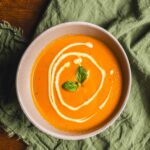 Creamy pumpkin soup garnished with a drizzle of cream and fresh basil leaves on top, served in a white bowl for a comforting and flavorful fall or winter meal.