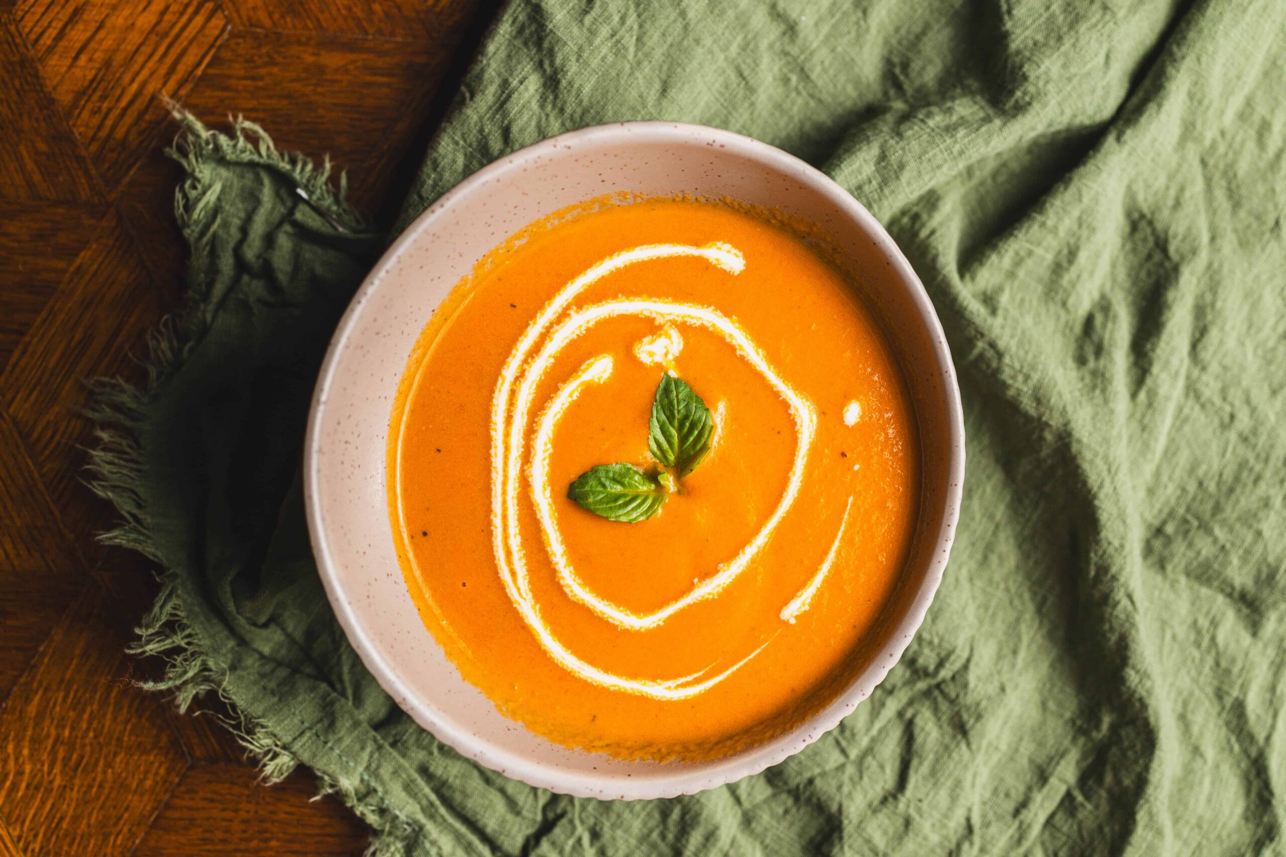 Creamy pumpkin soup garnished with a drizzle of cream and fresh basil leaves on top, served in a white bowl for a comforting and flavorful fall or winter meal.