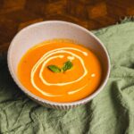 Creamy pumpkin soup garnished with a swirl of cream and fresh basil leaves, served in a ceramic bowl, on a rustic wooden table with a green cloth for a cozy and inviting presentation.