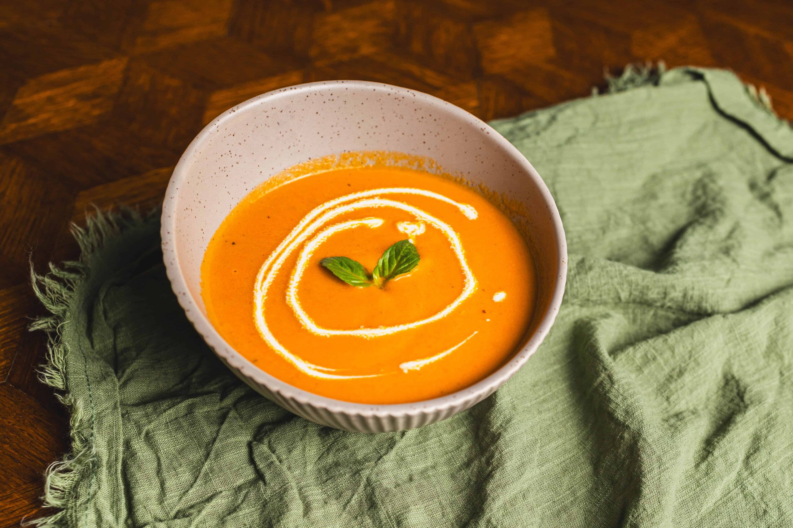 Creamy pumpkin soup garnished with a swirl of cream and fresh basil leaves, served in a ceramic bowl, on a rustic wooden table with a green cloth for a cozy and inviting presentation.