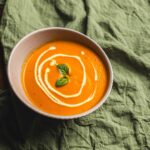 Creamy butternut squash soup garnished with fresh basil leaves and a swirl of cream, served in a rustic bowl on a wooden table with a green linen cloth.