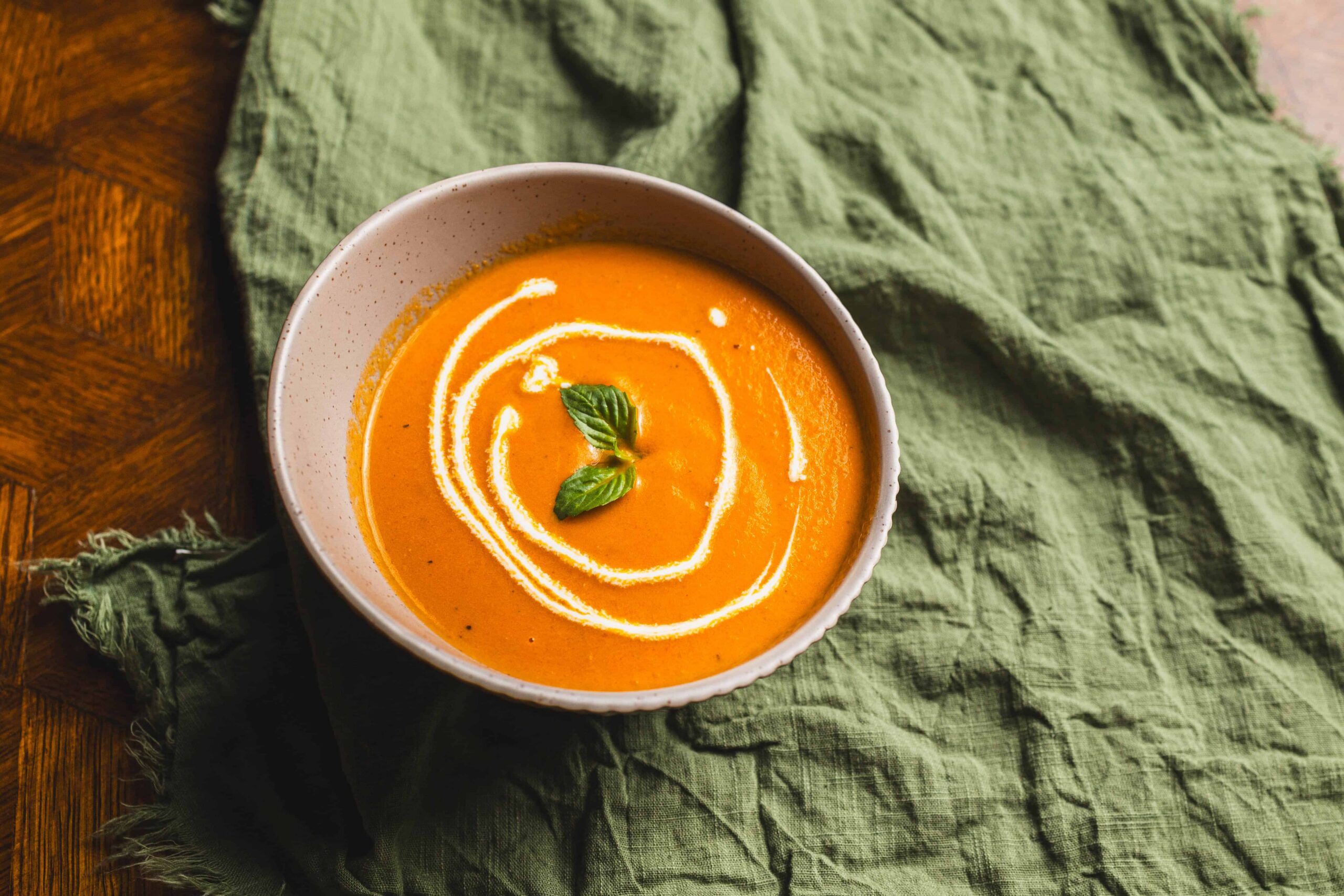 Creamy butternut squash soup garnished with fresh basil leaves and a swirl of cream, served in a rustic bowl on a wooden table with a green linen cloth.