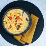 Creamy pasta with bell peppers and spinach in a white sauce, served in a black bowl on a black textured placemat with a mustard-colored napkin.