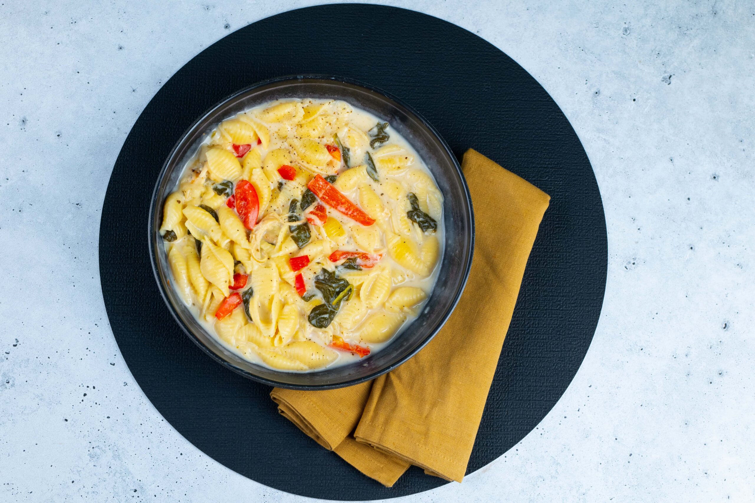 Creamy pasta with bell peppers and spinach in a white sauce, served in a black bowl on a black textured placemat with a mustard-colored napkin.