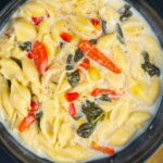 Creamy vegetable pasta with red bell peppers, spinach, and shells in a rich white sauce, served in a black bowl on a dark surface with a mustard yellow napkin.