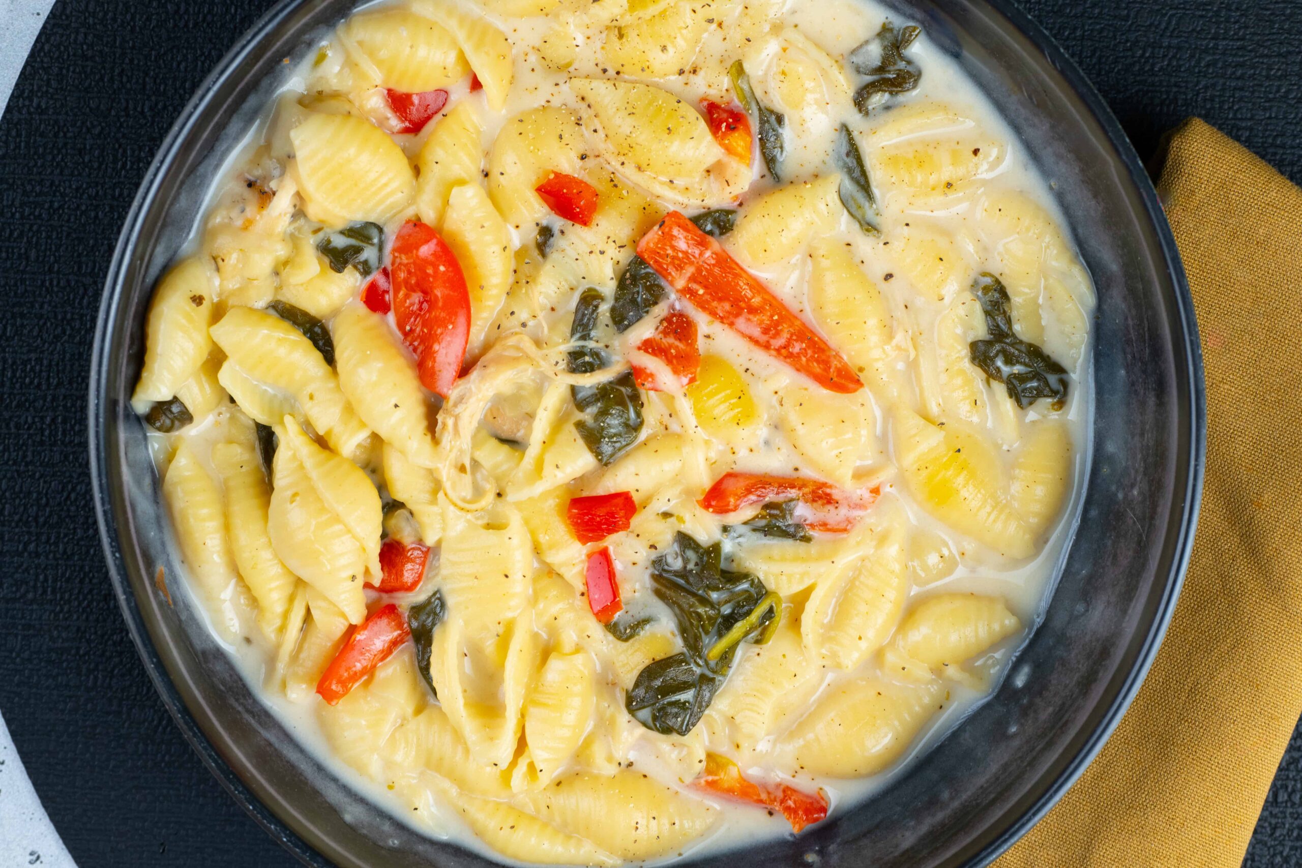 Creamy vegetable pasta with red bell peppers, spinach, and shells in a rich white sauce, served in a black bowl on a dark surface with a mustard yellow napkin.
