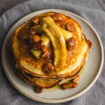 Golden brown pancakes topped with caramelized bananas, pecans, and syrup on a ceramic plate. Perfect for breakfast or brunch, these delicious pancakes are drizzled with syrup and packed with flavorful toppings.