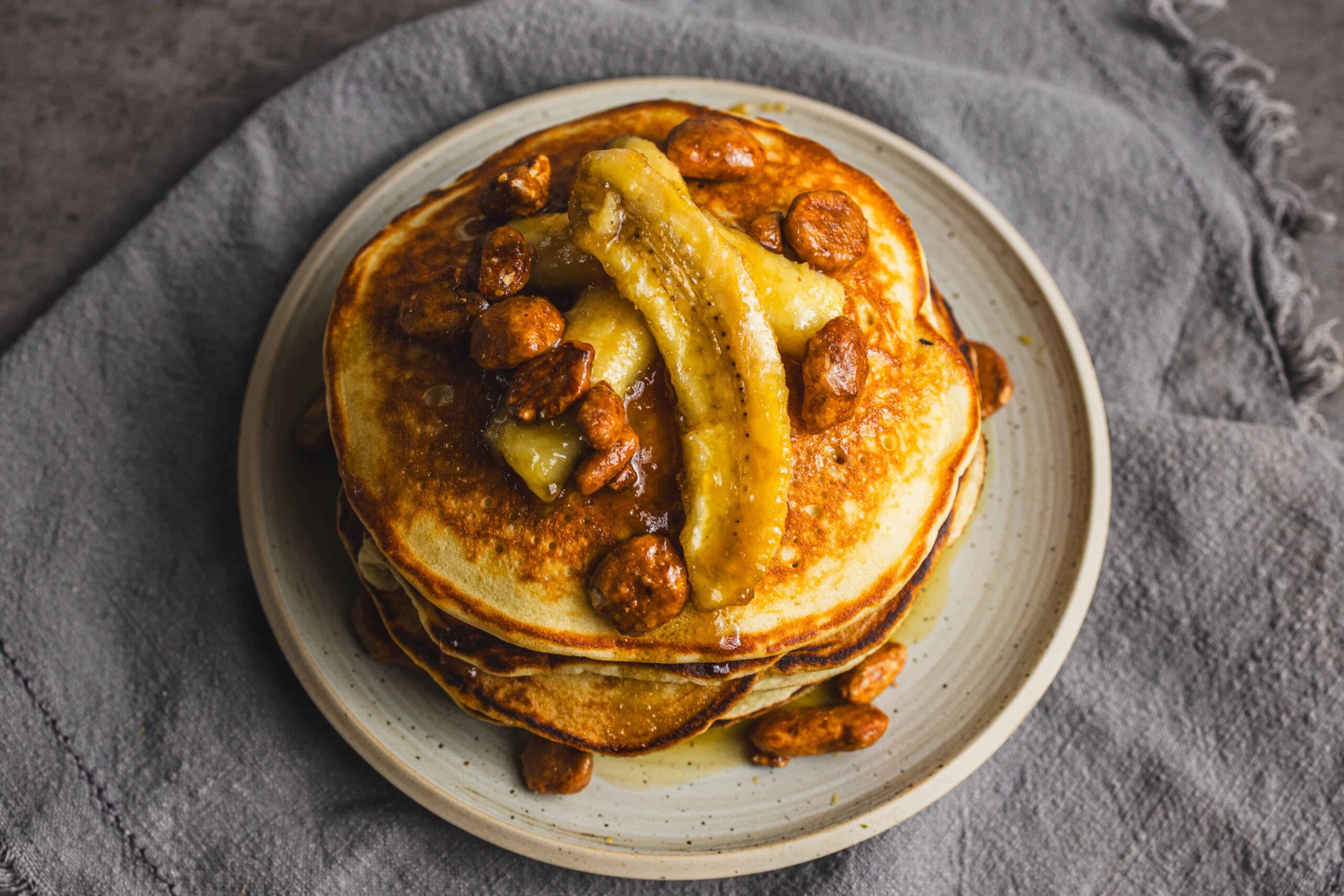 Golden brown pancakes topped with caramelized bananas, pecans, and syrup on a ceramic plate. Perfect for breakfast or brunch, these delicious pancakes are drizzled with syrup and packed with flavorful toppings.
