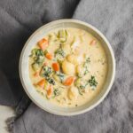 Creamy vegetable chicken stew in a white bowl on gray textured background, with carrots, potatoes, celery, and herbs, showcasing a comforting homemade soup recipe.