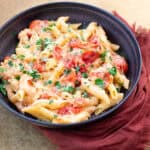 Creamy tomato and cheese pasta dish garnished with chopped parsley served in a black bowl on a textured surface with a red cloth napkin.
