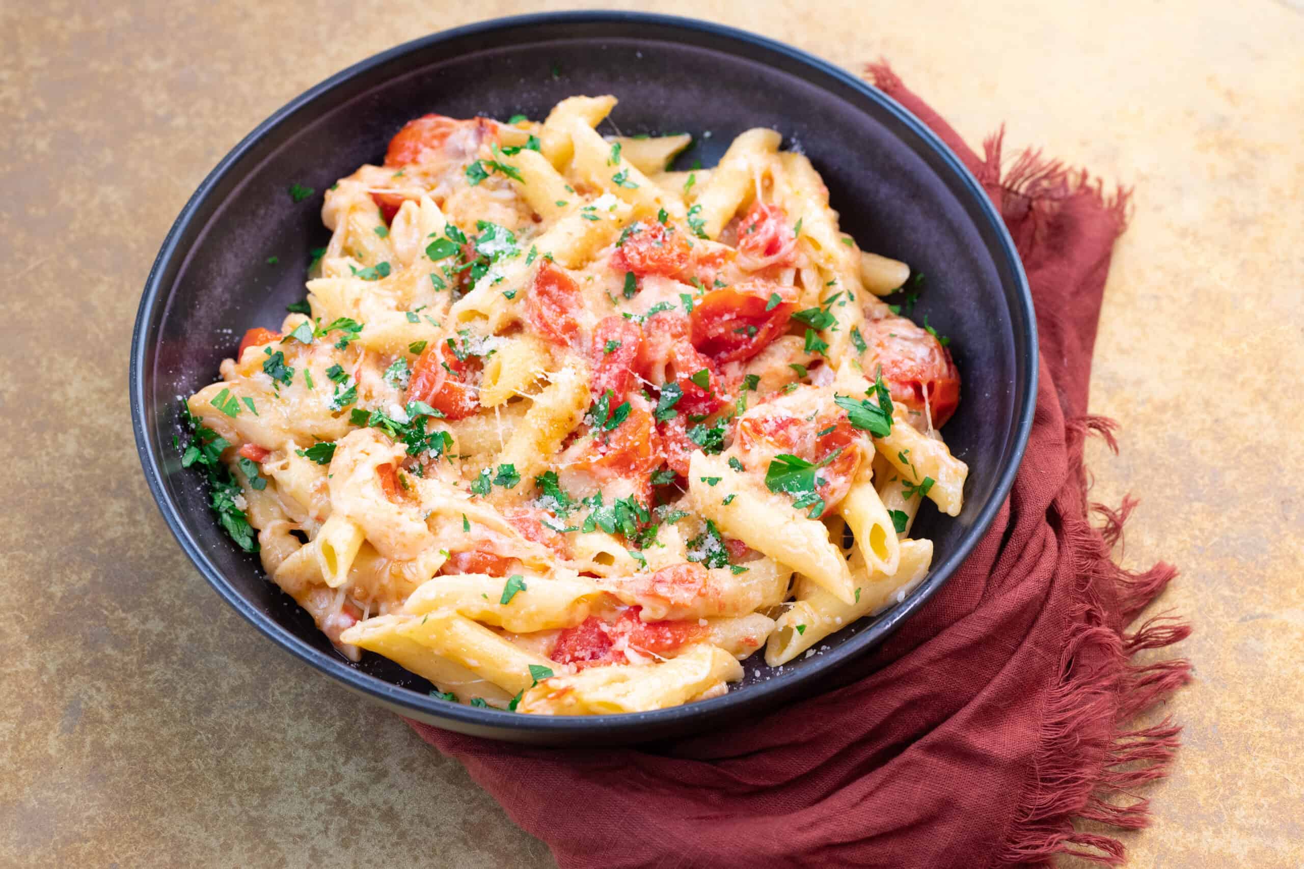 Creamy tomato and cheese pasta dish garnished with chopped parsley served in a black bowl on a textured surface with a red cloth napkin.