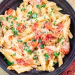 Creamy tomato and chicken pasta in a black bowl garnished with fresh parsley, served on a rustic beige surface with a red napkin for a cozy meal presentation.
