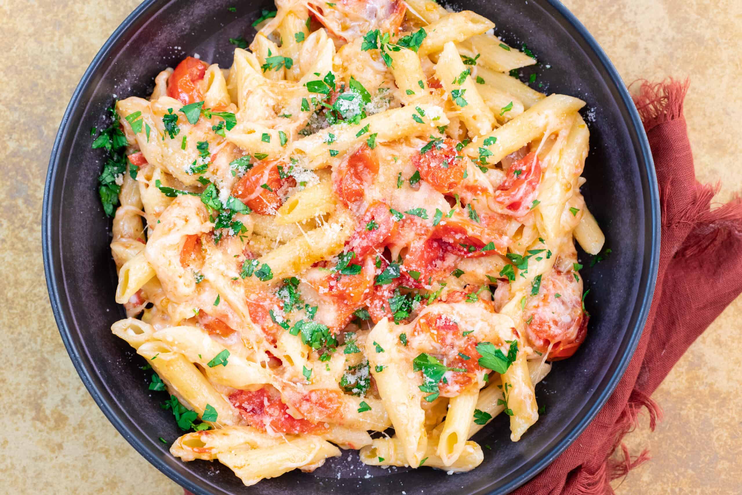Creamy tomato and chicken pasta in a black bowl garnished with fresh parsley, served on a rustic beige surface with a red napkin for a cozy meal presentation.