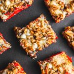 Buttered nut-topped brownies with a red velvet base, topped with chopped nuts, chocolate chips, and creamy frosting on a dark background.