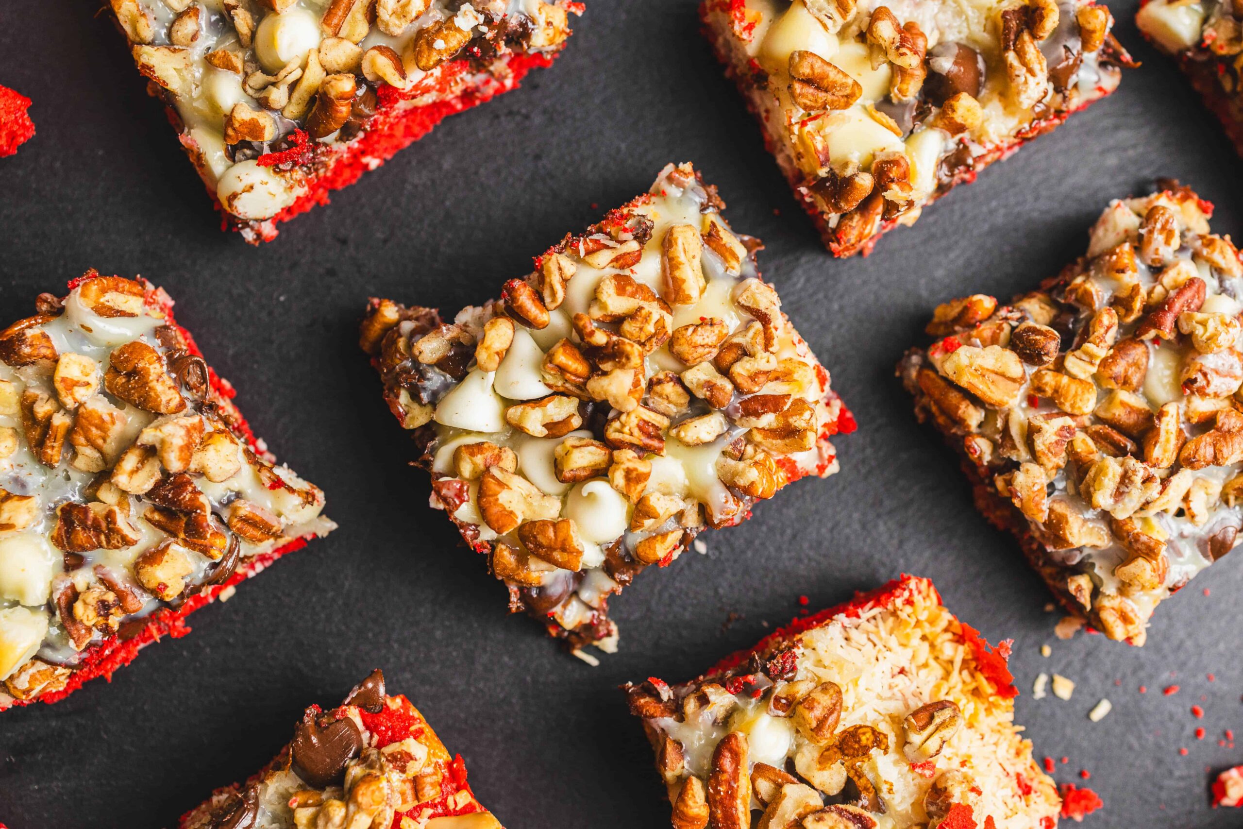Buttered nut-topped brownies with a red velvet base, topped with chopped nuts, chocolate chips, and creamy frosting on a dark background.
