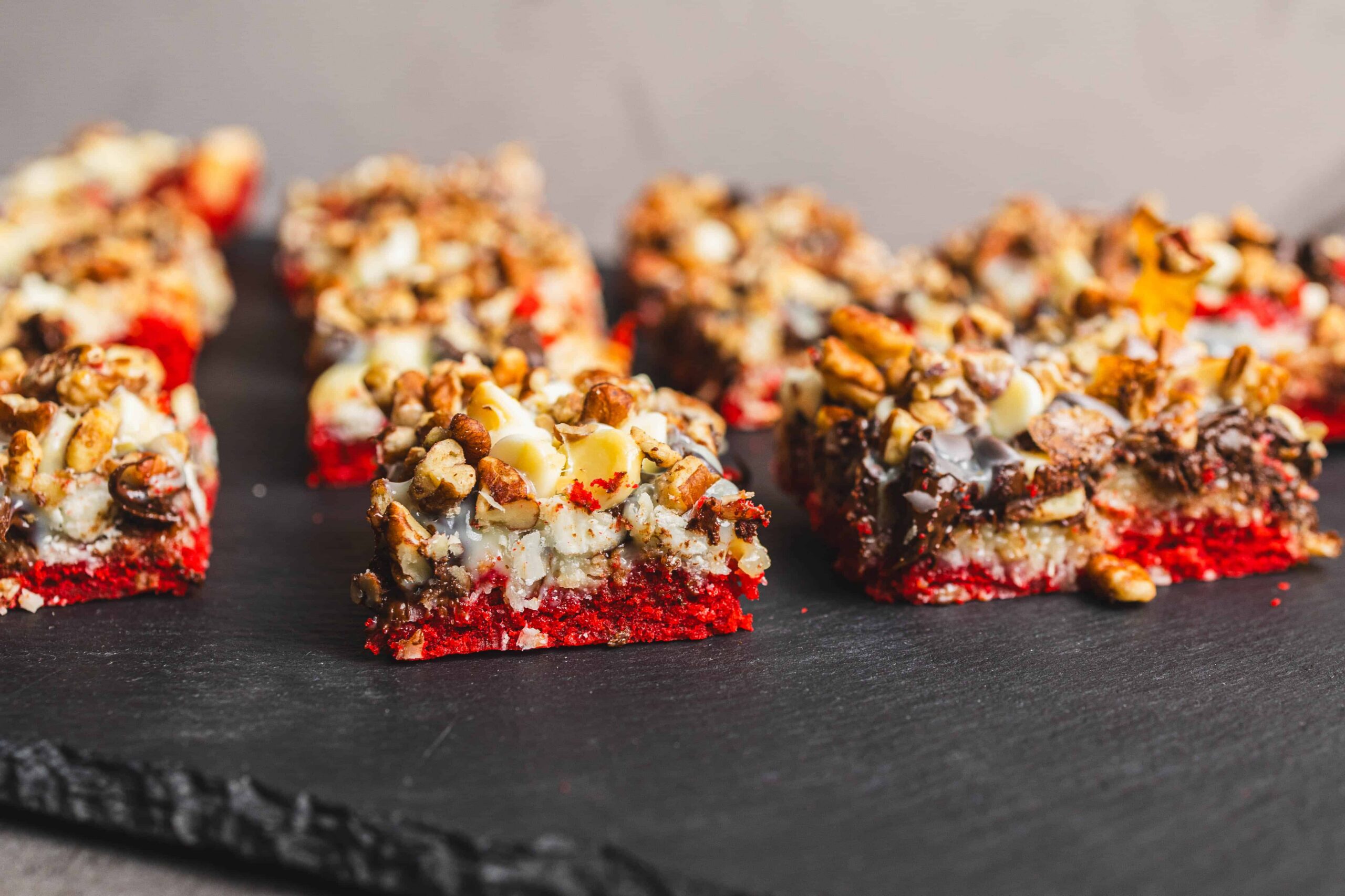 Red velvet no-bake dessert bars topped with chopped nuts and white chocolate on a black slate serving board.