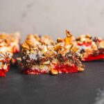Creamy fruit and nut rocky road dessert bars with red berry base, topped with chocolate, chopped nuts, and marshmallows on a dark slate surface. Perfect for holiday snacking and sweet treats.