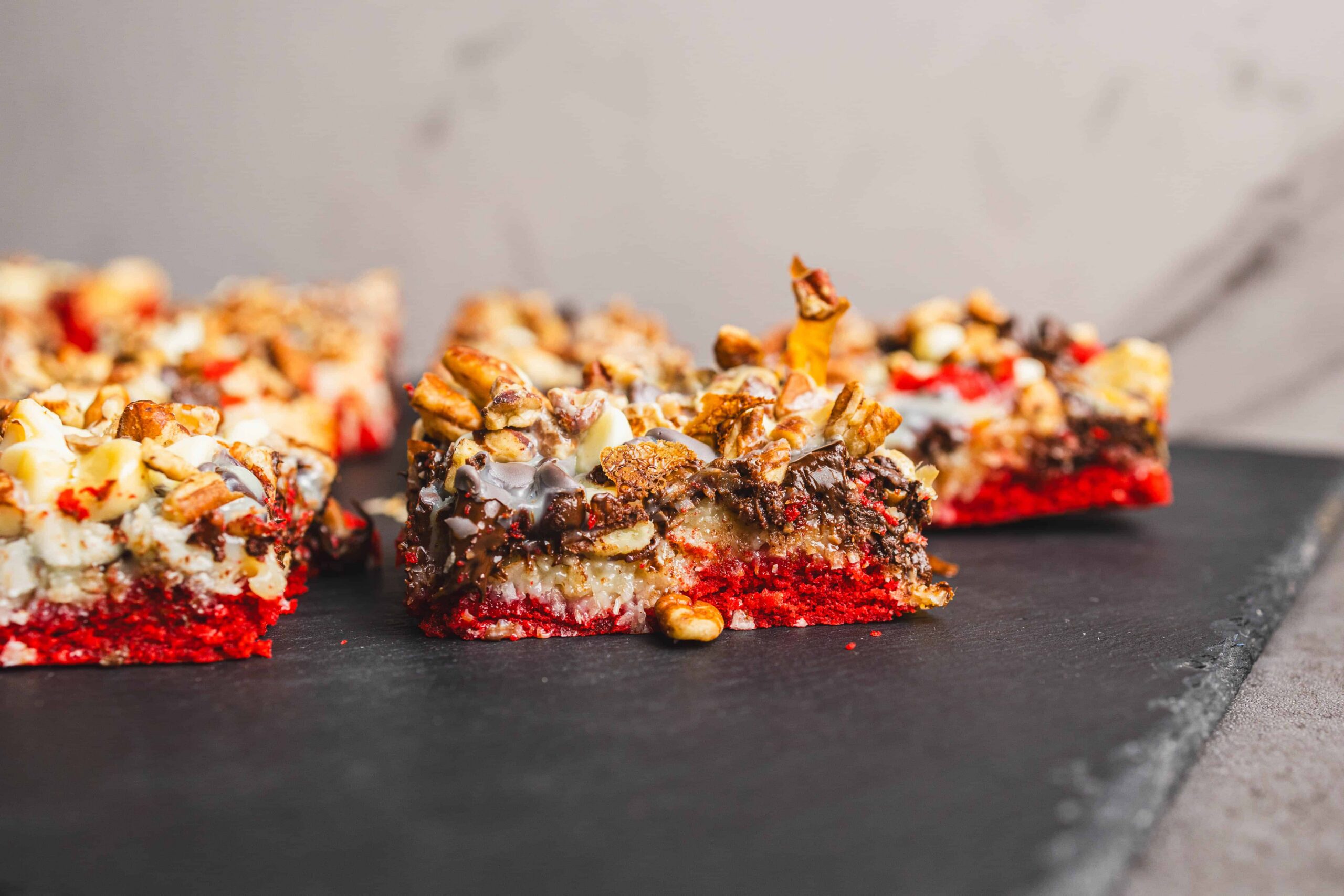 Creamy fruit and nut rocky road dessert bars with red berry base, topped with chocolate, chopped nuts, and marshmallows on a dark slate surface. Perfect for holiday snacking and sweet treats.