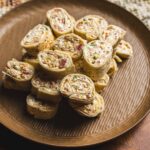 Savory rolled wrap sandwich slices filled with deli meats, cheeses, and vegetables on a brown textured plate.