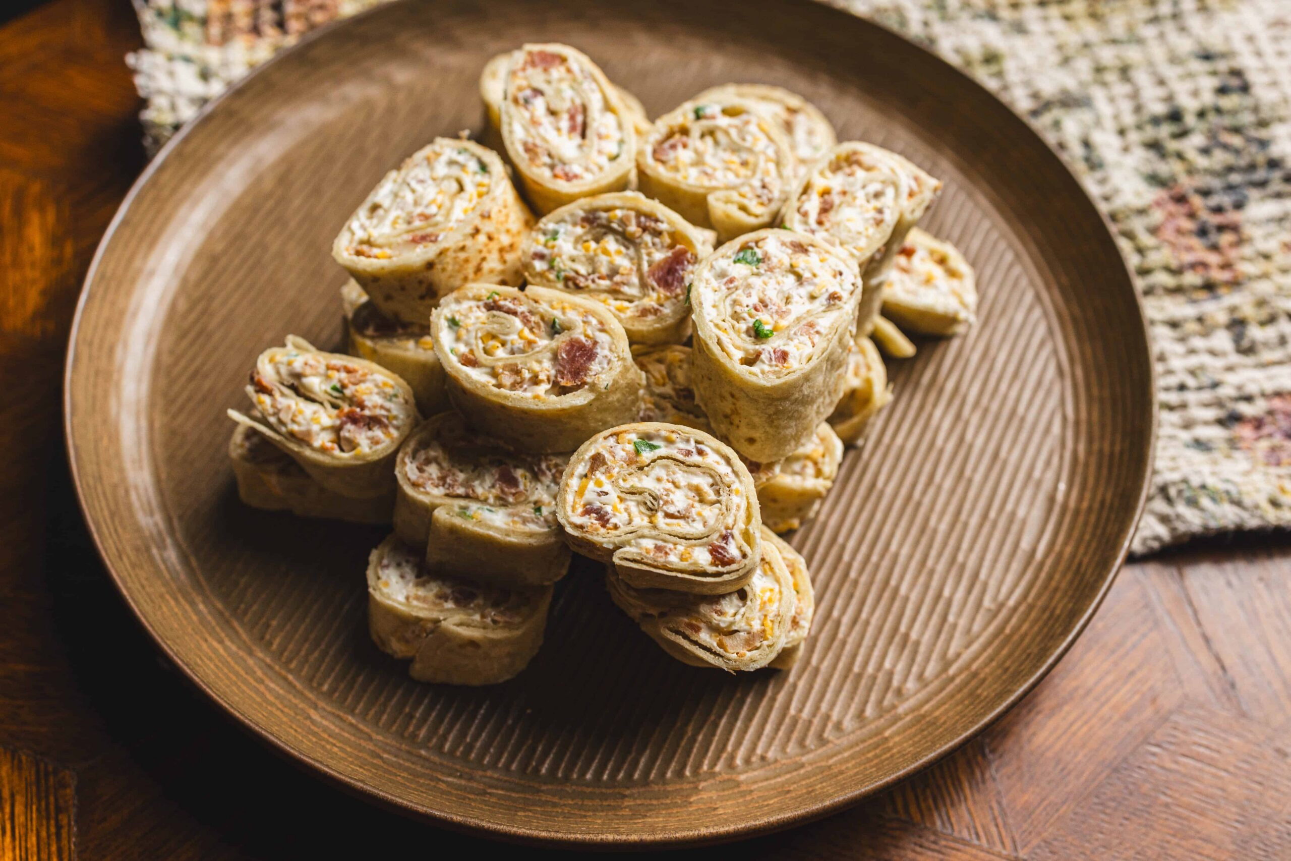 Savory rolled wrap sandwich slices filled with deli meats, cheeses, and vegetables on a brown textured plate.
