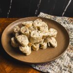 Creamy chicken salad wraps on a brown plate with chopped vegetables and herbs, served on a wooden table with a woven cloth, featuring fresh ingredients and appetizer presentation for party or snack ideas.