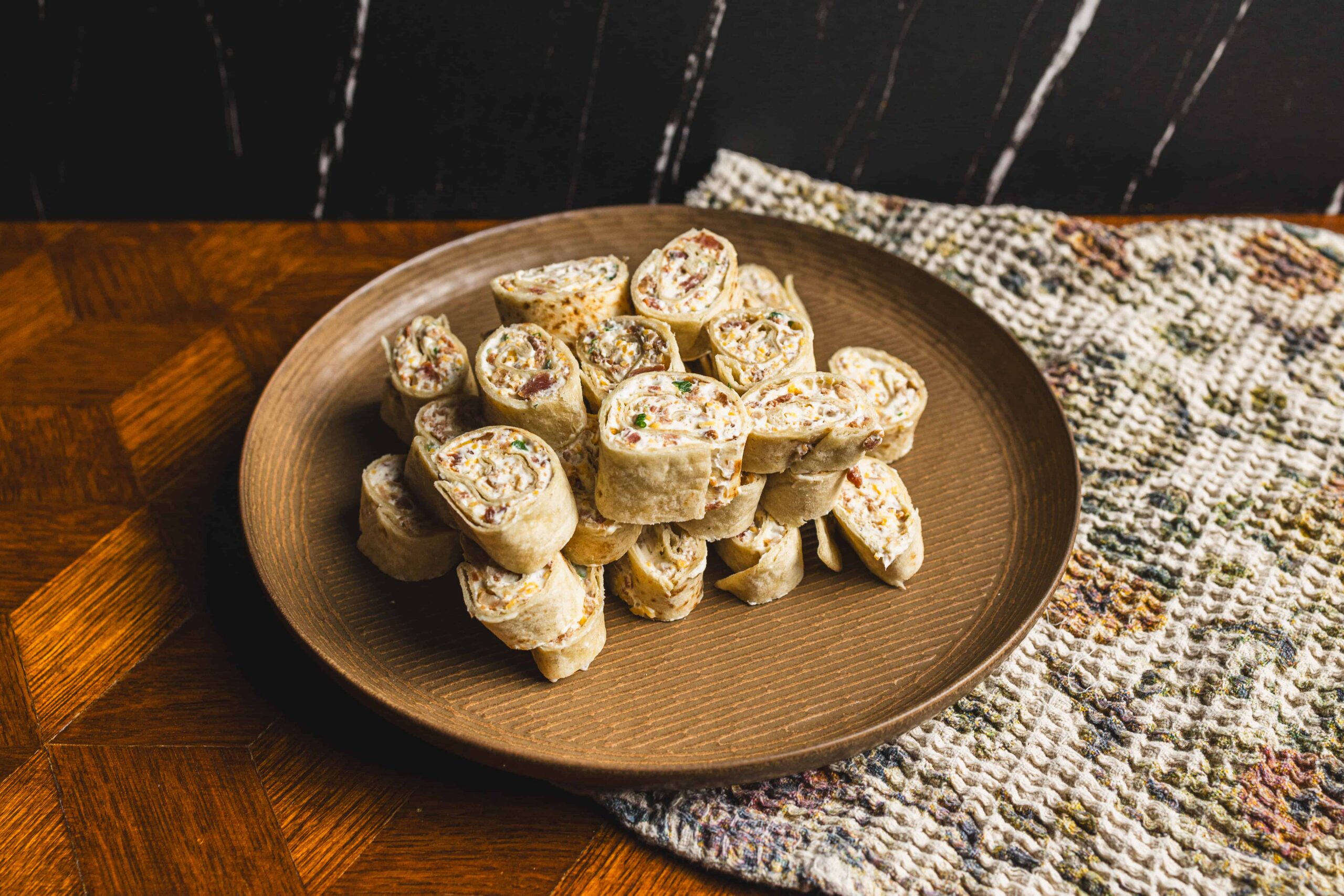 Creamy chicken salad wraps on a brown plate with chopped vegetables and herbs, served on a wooden table with a woven cloth, featuring fresh ingredients and appetizer presentation for party or snack ideas.